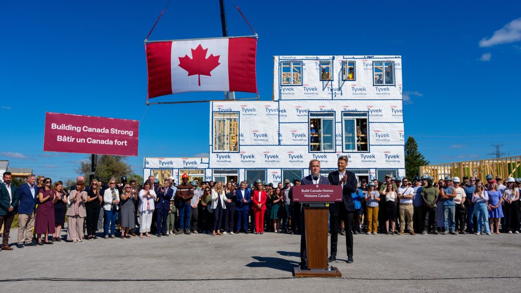 A crowd at an outdoor announcement in front of a modular house section and a large Canadian flag