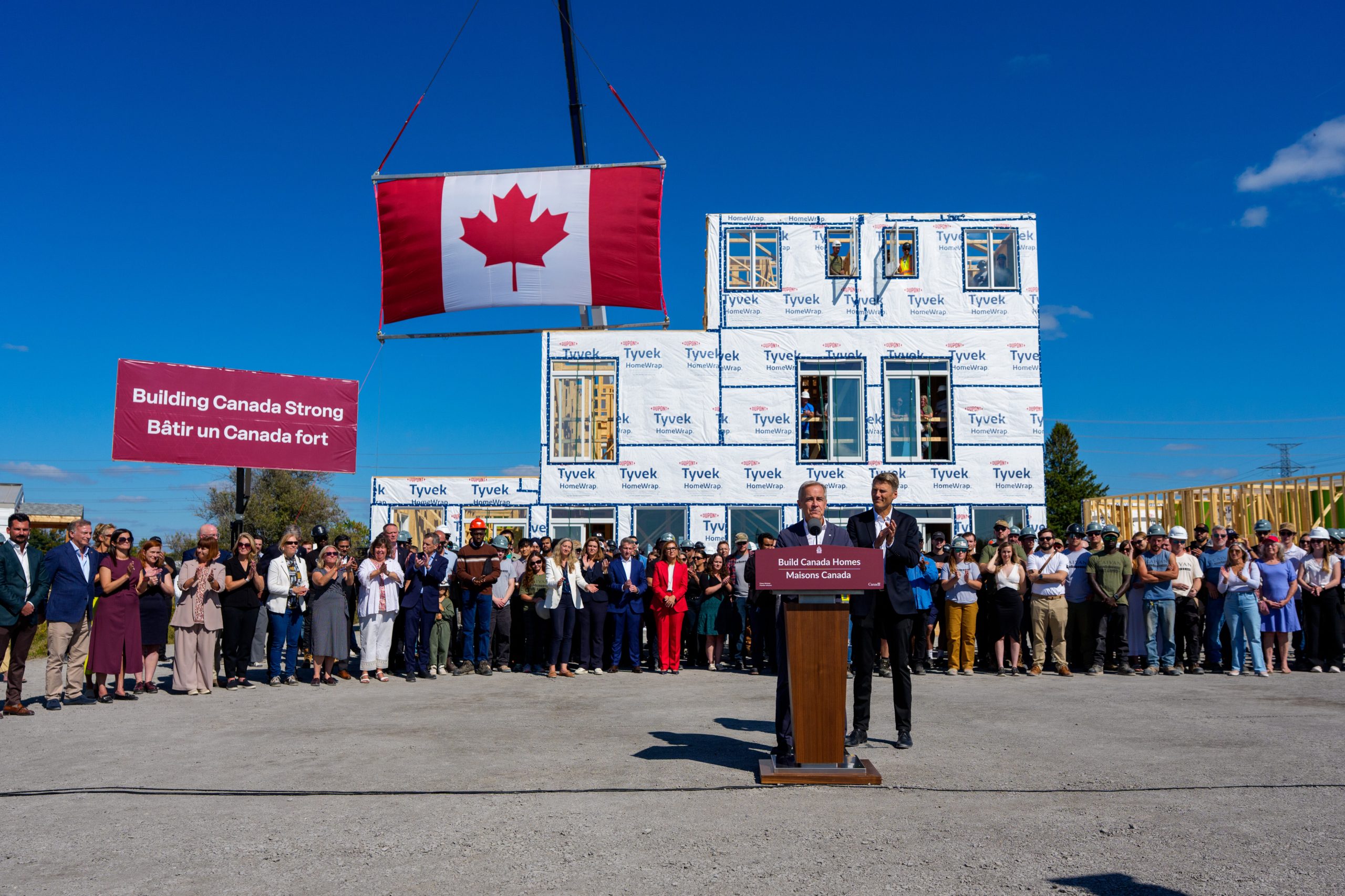 A crowd at an outdoor announcement in front of a modular house section and a large Canadian flag