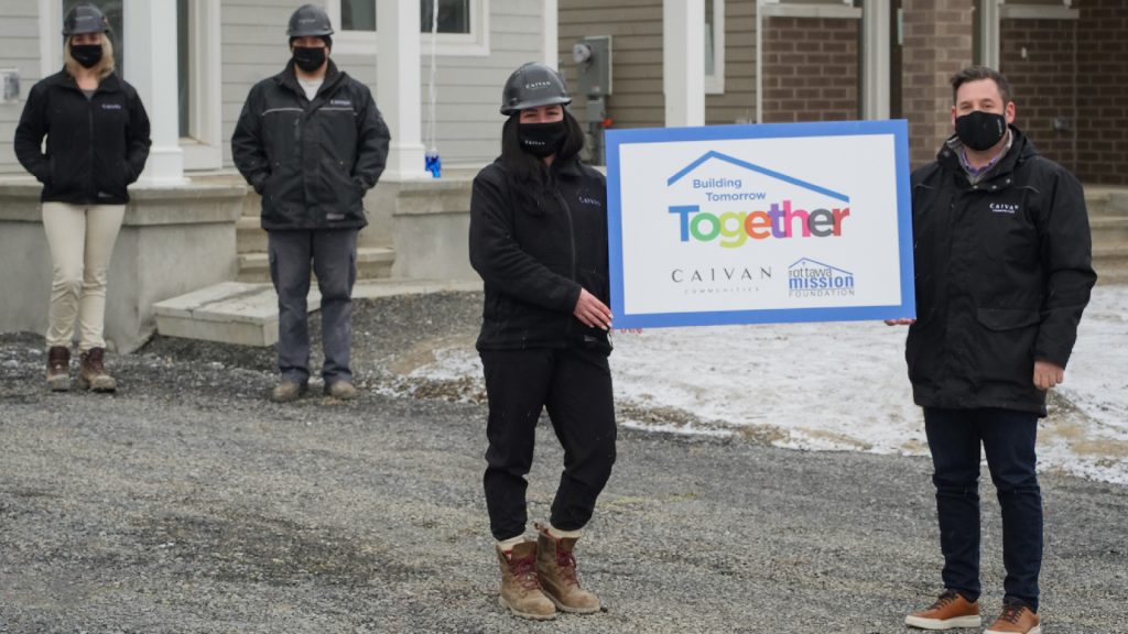 Three people outside a home under construction holding a Building Tomorrow Together sign