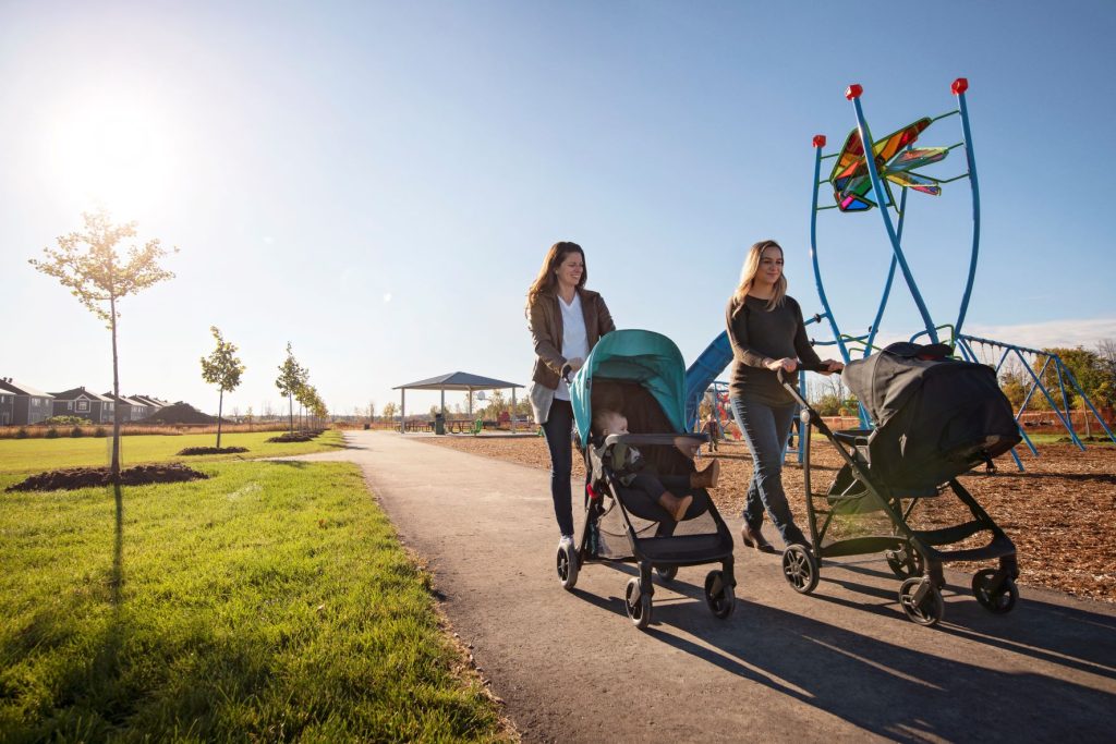 Two people pushing strollers along a park path past a climbing frame